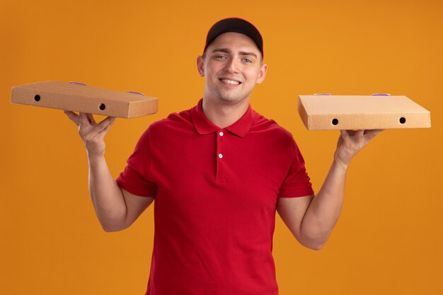 Souriant jeune livreur en uniforme avec capuchon tenant des boîtes de pizza isolé sur mur orange