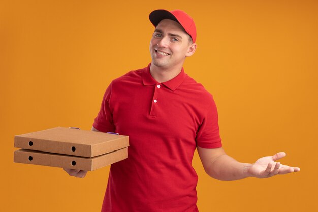 Souriant jeune livreur en uniforme avec capuchon tenant des boîtes de pizza propagation main isolé sur mur orange