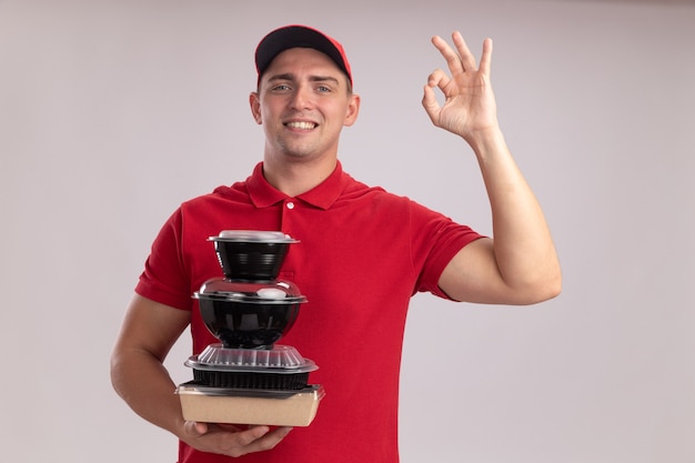 Souriant jeune livreur en uniforme avec capuchon tenant des contenants de nourriture montrant un geste correct isolé sur un mur blanc