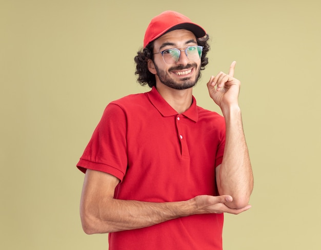 Souriant jeune livreur en uniforme rouge et casquette portant des lunettes regardant l'avant pointant vers le haut isolé sur un mur vert olive