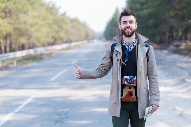 Souriant jeune mâle touristique avec appareil photo vintage autour du cou, faisant de l&#39;auto-stop le long d&#39;une route