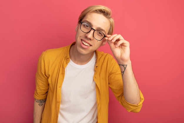 Souriant jeune mec blond portant un t-shirt jaune et des lunettes et a attrapé des lunettes isolées sur rose