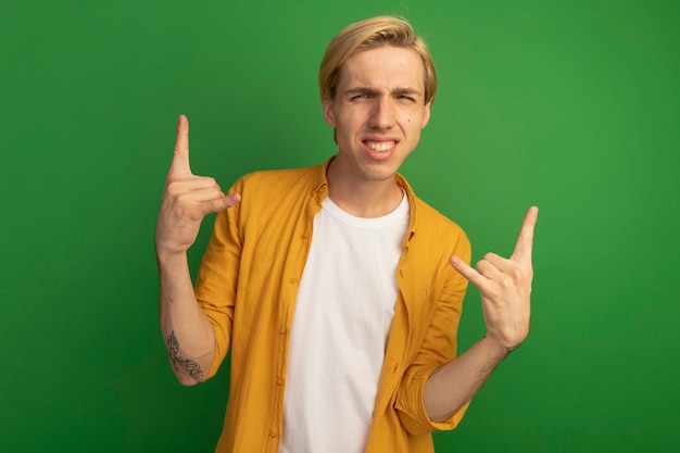 Souriant jeune mec blond portant un t-shirt jaune montrant le geste de la chèvre