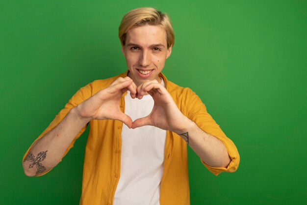 Souriant jeune mec blond portant un t-shirt jaune montrant le geste du cœur isolé sur vert