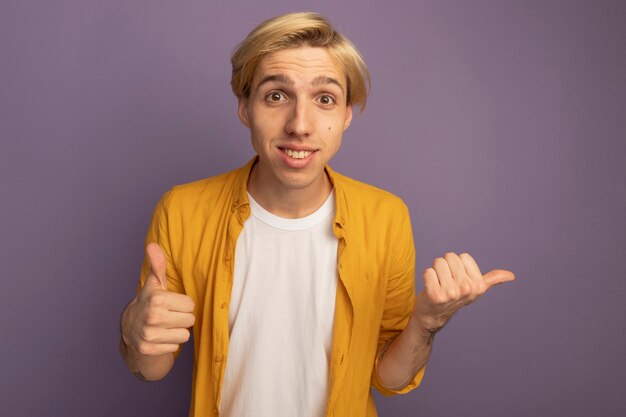 Souriant jeune mec blond portant un t-shirt jaune montrant le pouce vers le haut isolé sur violet