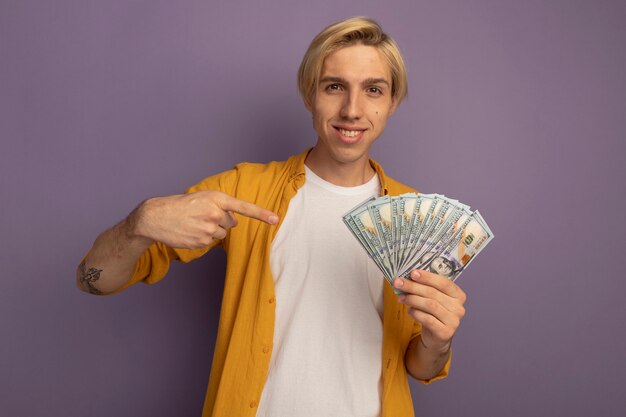 Souriant jeune mec blond portant un t-shirt jaune tenant et points à l'argent isolé sur violet