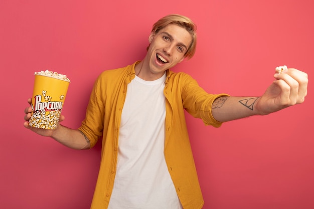 Souriant jeune mec blond portant un t-shirt jaune tenant un seau de pop-corn et tenant un morceau de pop-corn isolé sur rose