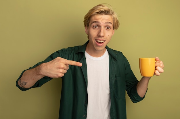 Souriant jeune mec blond portant un t-shirt vert tenant et pointe une tasse de thé