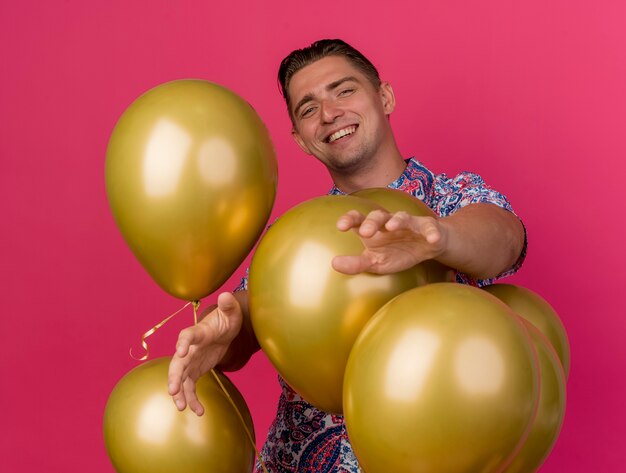 Souriant jeune mec de fête portant une chemise colorée debout derrière des ballons tenant les mains isolés sur rose