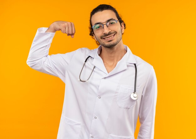 Souriant jeune médecin avec des lunettes médicales portant une robe médicale avec stéthoscope faisant un geste fort sur jaune