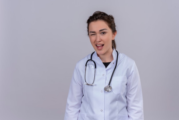 Souriant jeune médecin portant une blouse médicale portant un stéthoscope cligna des yeux sur le mur blanc