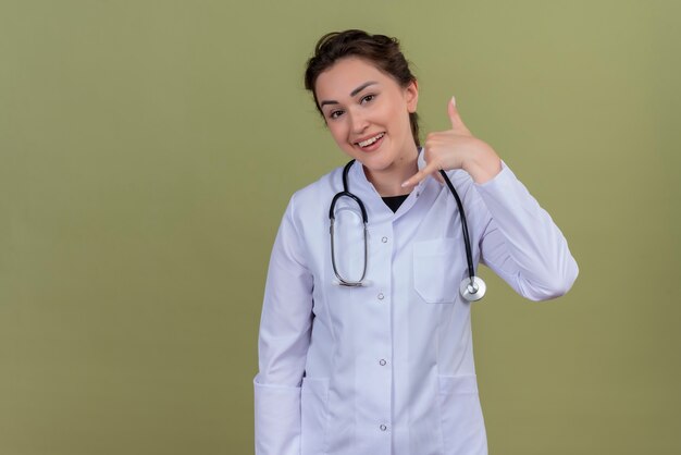 Souriant jeune médecin portant une blouse médicale portant un stéthoscope montre un appel téléphonique sur le mur vert