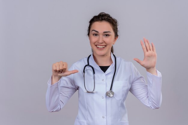 Souriant jeune médecin portant une blouse médicale portant un stéthoscope montre un geste différent sur le mur blanc