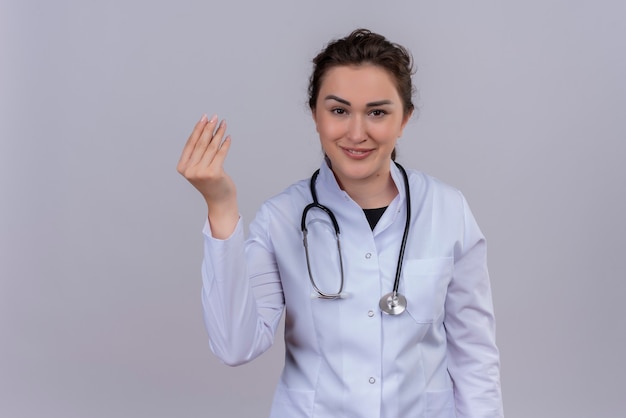 Souriant jeune médecin portant une blouse médicale portant un stéthoscope montre un geste de trésorerie sur un mur blanc