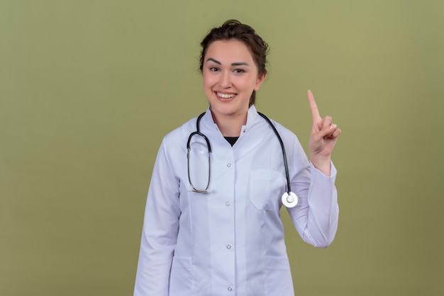 Souriant jeune médecin portant une blouse médicale portant un stéthoscope montre un sur le mur vert