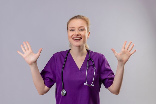 Souriant jeune médecin portant une blouse médicale violette et un stéthoscope se propage les mains sur un mur blanc isolé