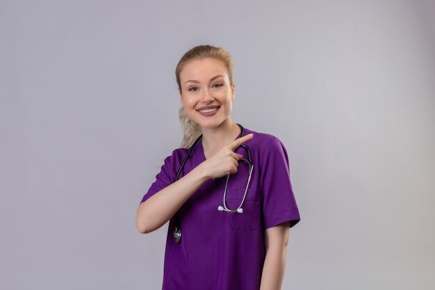 Souriant jeune médecin portant une robe médicale violette et un stéthoscope pointe vers le côté sur un mur blanc isolé