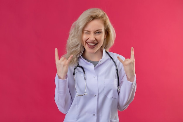 Souriant jeune médecin portant un stéthoscope en blouse médicale cligna des yeux montrant le geste de chèvre sur fond rouge