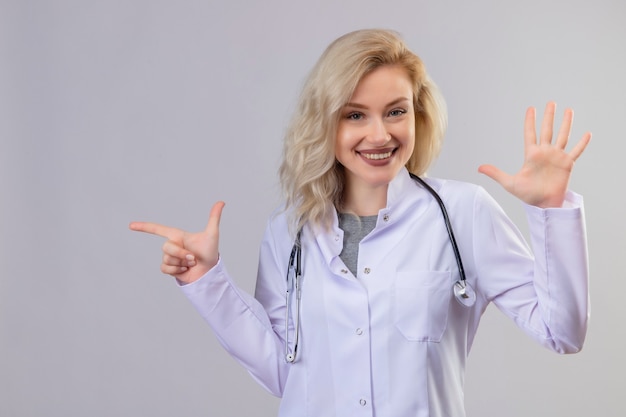 Souriant jeune médecin portant un stéthoscope en robe médicale montrant un geste différent sur un mur blanc