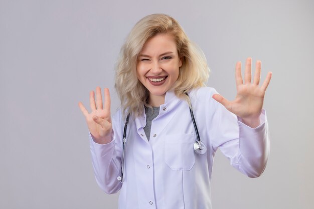 Souriant jeune médecin portant un stéthoscope en robe médicale montrant un geste différent sur un mur blanc