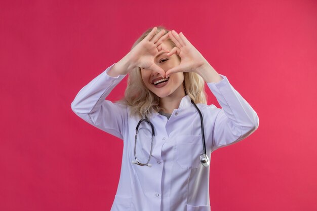 Souriant jeune médecin portant un stéthoscope en robe médicale montrant le geste du cœur sur fond rouge