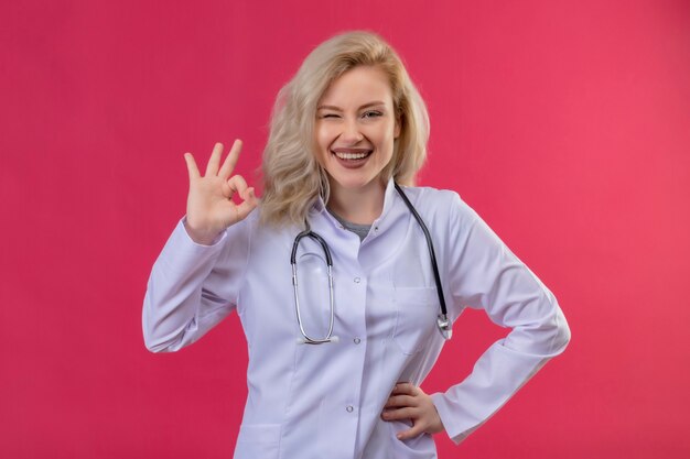 Souriant jeune médecin portant un stéthoscope en robe médicale montrant le geste okey sur fond rouge