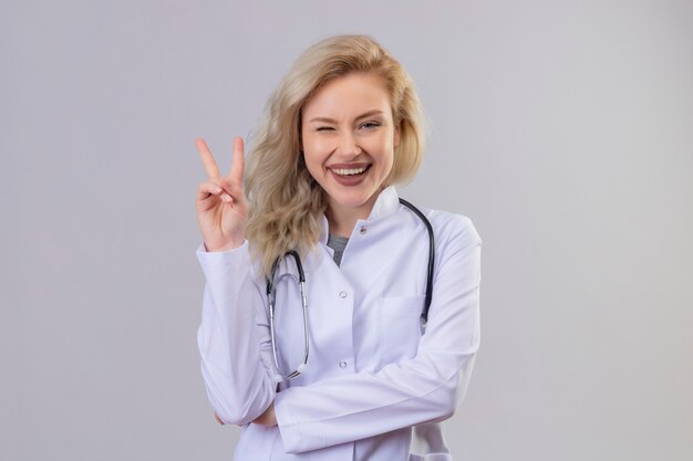 Souriant jeune médecin portant un stéthoscope en robe médicale montrant le geste de paix sur le mur blanc