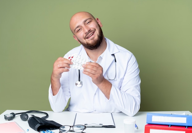 Photo gratuite souriant jeune médecin de sexe masculin chauve portant une robe médicale et un stéthoscope assis au travail de bureau avec des outils médicaux tenant des pilules isolées sur le mur vert