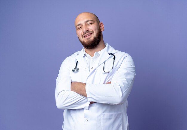 Souriant jeune médecin de sexe masculin chauve portant une robe médicale et un stéthoscope croisant les mains isolés sur fond bleu