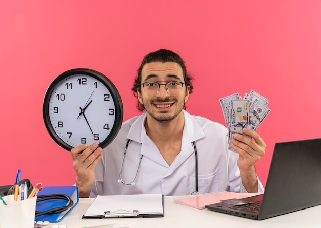 Souriant jeune médecin de sexe masculin avec des lunettes médicales portant une robe médicale avec stéthoscope assis au bureau