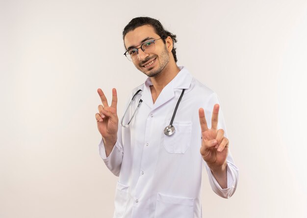 Souriant jeune médecin de sexe masculin avec des lunettes optiques portant une robe blanche avec stéthoscope montrant le geste de paix sur un mur blanc isolé avec espace de copie