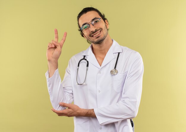 Souriant jeune médecin de sexe masculin avec des lunettes optiques portant une robe blanche avec stéthoscope montrant le geste de paix sur le vert