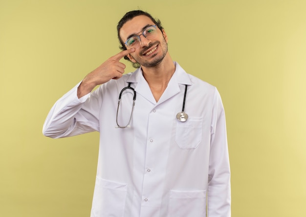 Souriant jeune médecin de sexe masculin avec des lunettes optiques portant une robe blanche avec stéthoscope pointe son œil sur le vert