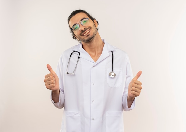 Souriant jeune médecin de sexe masculin avec des lunettes optiques portant une robe blanche avec stéthoscope ses pouces vers le haut sur un mur blanc isolé avec copie espace