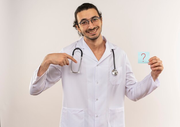 Souriant jeune médecin de sexe masculin avec des lunettes optiques portant une robe blanche avec stéthoscope tenant et pointe vers le point d'interrogation papier sur un mur blanc isolé avec copie espace