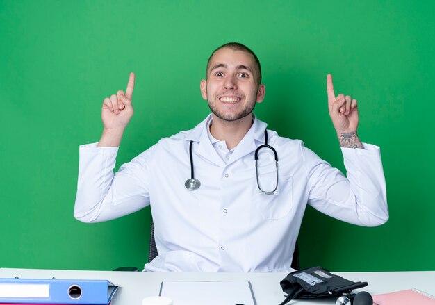 Souriant jeune médecin de sexe masculin portant une robe médicale et un stéthoscope assis au bureau avec des outils de travail pointant vers le haut avec les doigts isolés sur le mur vert