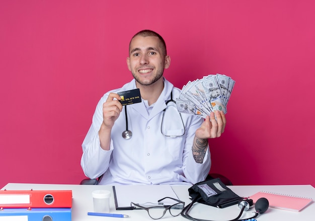 Photo gratuite souriant jeune médecin de sexe masculin portant une robe médicale et un stéthoscope assis au bureau avec des outils de travail tenant de l'argent et une carte de crédit isolé sur un mur rose
