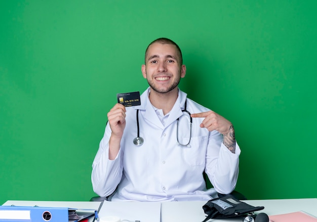 Souriant jeune médecin de sexe masculin portant une robe médicale et un stéthoscope assis au bureau avec des outils de travail tenant et pointant sur la carte de crédit isolée sur le mur vert