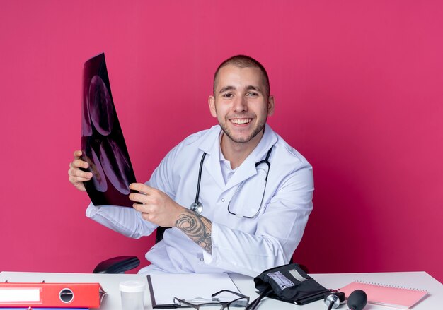 Souriant jeune médecin de sexe masculin portant une robe médicale et un stéthoscope assis au bureau avec des outils de travail tenant un tir aux rayons x isolé sur un mur rose