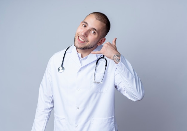 Souriant jeune médecin de sexe masculin portant une robe médicale et un stéthoscope autour de son cou faisant appel geste isolé sur mur blanc