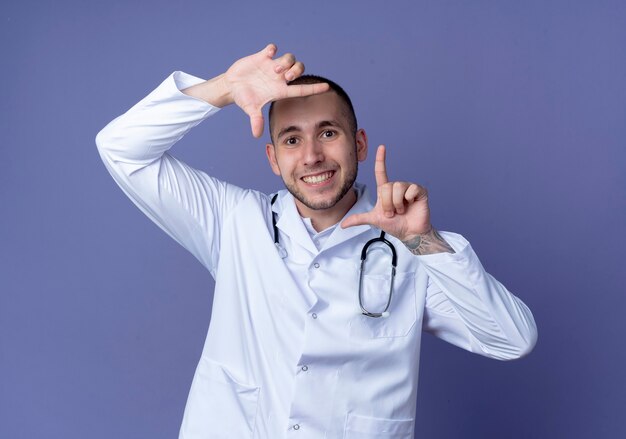 Souriant jeune médecin de sexe masculin portant une robe médicale et un stéthoscope autour de son cou faisant le geste du cadre isolé sur le mur violet