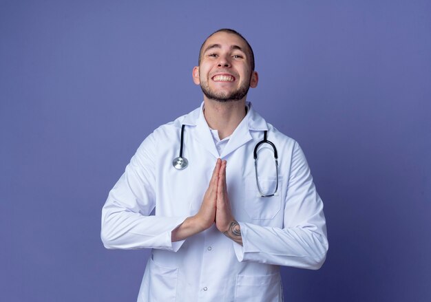 Souriant jeune médecin de sexe masculin portant une robe médicale et un stéthoscope autour de son cou mettant les mains dans le geste de prier isolé sur le mur violet