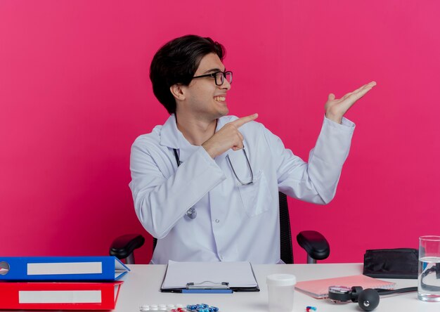 Souriant jeune médecin de sexe masculin portant une robe médicale et un stéthoscope avec des lunettes assis au bureau avec des outils médicaux tournant la tête à l'autre montrant la main vide pointant sur le côté isolé sur le mur rose