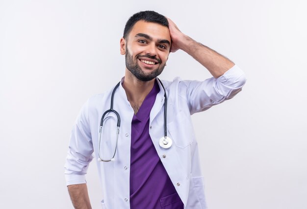 Souriant jeune médecin de sexe masculin portant une robe médicale stéthoscope a mis sa main sur la tête sur fond blanc isolé