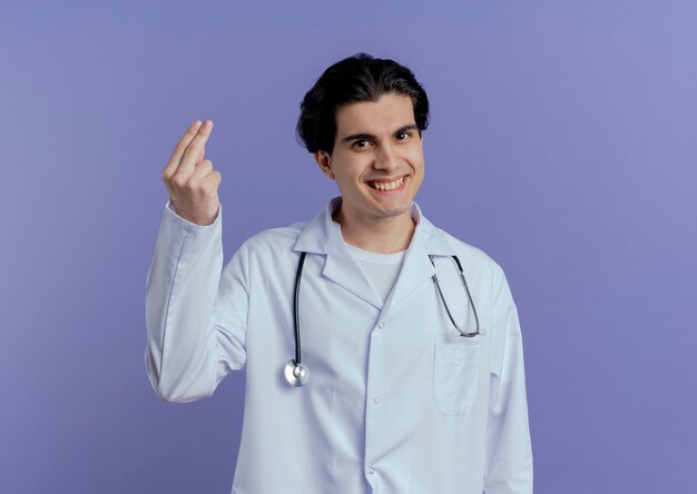 Souriant jeune médecin de sexe masculin portant une robe médicale et un stéthoscope montrant deux avec la main isolée sur un mur violet avec espace de copie