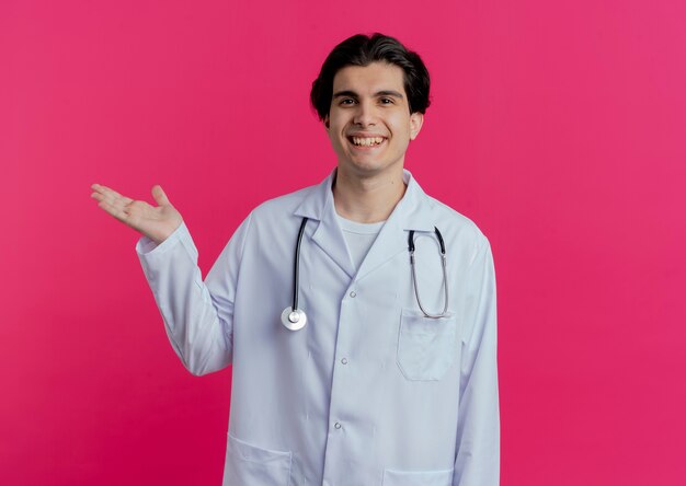 Souriant jeune médecin de sexe masculin portant une robe médicale et un stéthoscope montrant une main vide isolée sur un mur rose avec espace de copie