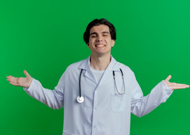 Souriant jeune médecin de sexe masculin portant une robe médicale et un stéthoscope montrant les mains vides avec les yeux fermés isolés sur le mur vert