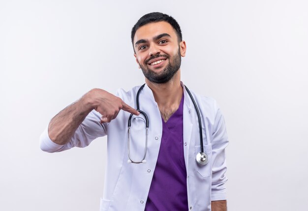 Souriant jeune médecin de sexe masculin portant une robe médicale stéthoscope pointe vers lui-même sur fond blanc isolé