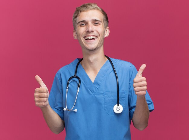 Souriant jeune médecin de sexe masculin portant un uniforme de médecin avec stéthoscope montrant les pouces vers le haut isolé sur mur rose