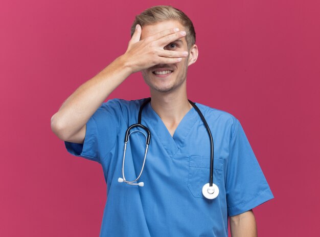 Souriant jeune médecin de sexe masculin portant un uniforme de médecin avec un visage couvert de stéthoscope avec une main isolée sur un mur rose